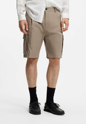 Mann trägt beige Cargoshorts, schwarze Socken und schwarze Leder-Mokassins, mit einem weißem Hemd, das teilweise in die Hose gesteckt ist.