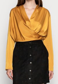 Satynowa, musztardowa bluzka z długim rękawem i marszczonym przodem, zestawiona z czarną zamszową spódnicą z pięcioma srebrnymi guzikami z przodu.