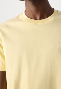 Primer plano de una persona que lleva una camiseta de cuello redondo de color amarillo claro con un texto blanco tenue en la parte superior derecha.
