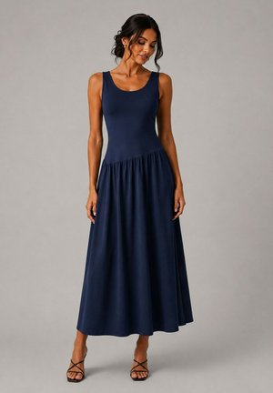 Femme en robe longue bleu marine sans manches avec un corsage ajusté et une jupe évasée, portant des sandales noires à talons à brides, debout sur un fond gris.