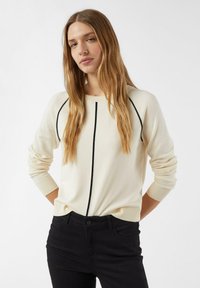 Frau mit langen blonden Haaren, die einen cremefarbenen Pullover mit schwarzen vertikalen und diagonalen Linien trägt, kombiniert mit einer schwarzen Hose, stehend vor einem weißen Hintergrund.