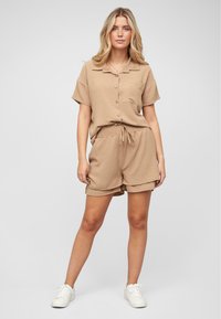 Ensemble comprenant une chemise à manches courtes boutonnée texturée de couleur beige et un short assorti. Taille élastique avec cordon de serrage, accompagné de baskets blanches.