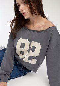 Szary sweatshirt z odkrytymi ramionami z dużą białą cyfrą "82" nadrukowaną z przodu, z długimi rękawami i luźnym krojem, w połączeniu z niebieskimi dżinsami.