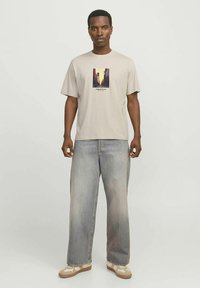 Beige t-paita, jossa on painettu kaupunkimaisemakuva ja teksti, yhdistettynä löysälahkeisiin haalistuneisiin harmaisiin farkkuihin ja kermanvärisiin lenkkareihin.