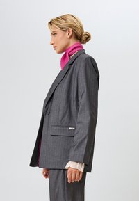 Harmaa raidallinen pikkutakki, jossa on viiltoapikaset, yksittäinen nappilukitus ja näkyvä vaaleanpunainen vuori, jota täydentää vaaleanpunainen huivi.