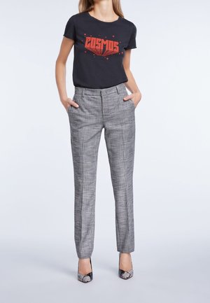 Zwarte t-shirt met rode "COSMOS" grafiek, gecombineerd met grijs geruite maatwerkbroek. Vrouw staat met haar handen in haar zakken, draagt gepatternde hakken.