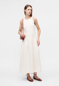 Μακρύ, αμάνικο κρεμ φόρεμα με ζώνη στη μέση και εμφανείς ραφές. Συνδυασμένο με καφέ, δερμάτινα παπούτσια με κορδόνια και μικρή κόκκινη τσάντα.