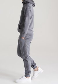 Šedý teplákový komplet s kapucí, který má podél boků texturovaný pruh. V kombinaci s bílými teniskami, které mají elegantní sportovní design.