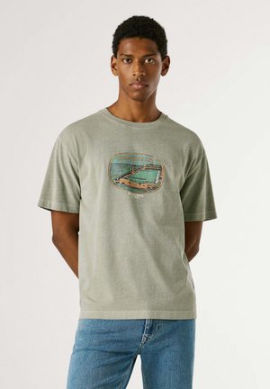 Jonge man in een lichtgroen T-shirt met Ramsgate Lido-graphic en blauwe jeans, staand met handen achter de rug, neutrale uitdrukking.