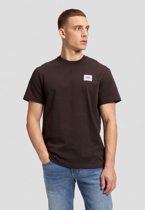 Homme portant un t-shirt marron foncé de la marque Lee à manches courtes et un jean bleu clair, debout avec une main dans la poche, regardant sur le côté.