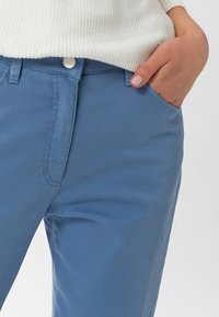 Henkilö, jolla on ylläsiniset housut ja valkoinen neulepaita, ja yksi käsi lepää housujen etutaskussa.