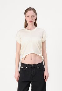 T-shirt cropped crema con maniche corte arrotolate, caratterizzato da lacci regolabili all'orlo. Abbinato a jeans neri a vita alta.