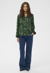 Green patterned long-sleeve blouse with a collar and button placket, paired with blue flared jeans and dark shoes. Smooth fabric and relaxed fit.