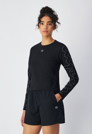 Schwarz langärmliges Shirt mit weißen Zickzacknähten an den Ärmeln, runder Ausschnitt und Logo auf der Brust, kombiniert mit schwarzen Shorts.