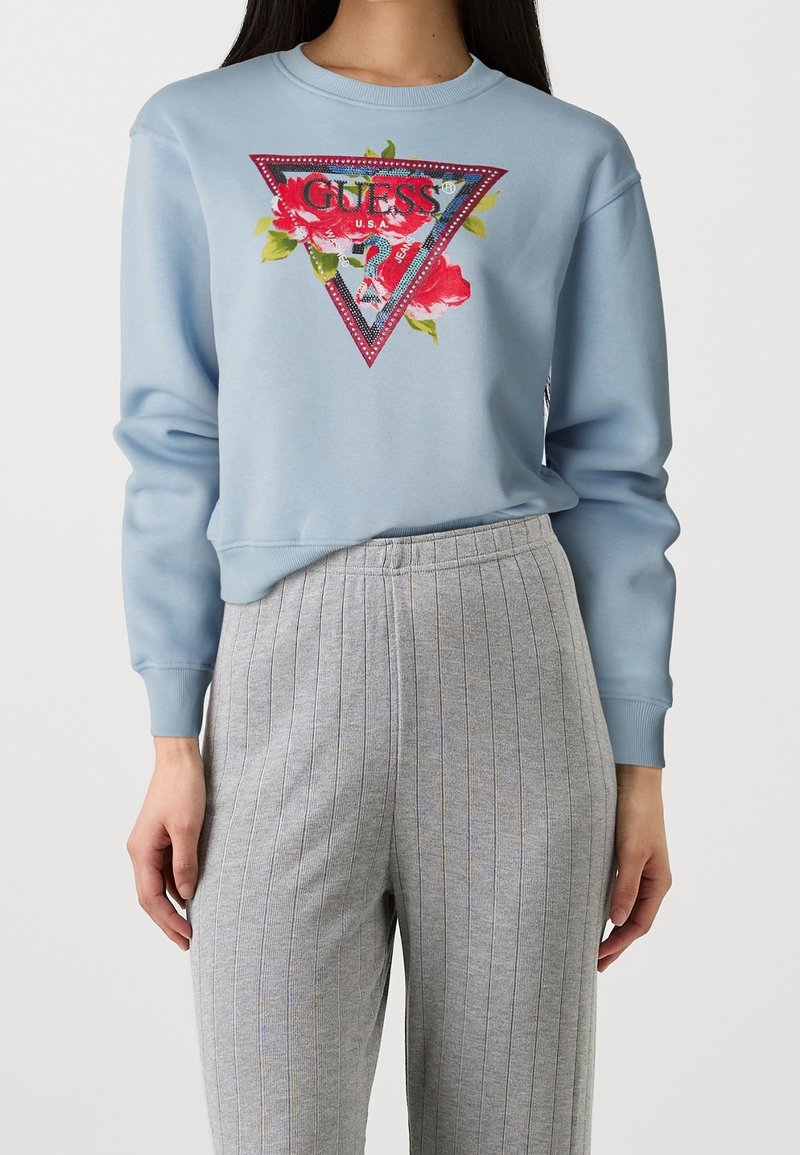 Femme portant un sweat-shirt bleu clair avec le logo floral rouge GUESS et un pantalon gris à fines rayures taille haute, debout devant un fond blanc.