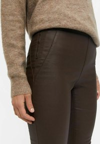 Lähikuva henkilöstä, joka käyttää ruskeita nahkahousuja, joissa on sivuvetoketjullinen tasku, ja näkyvissä on beigenvärisen neuleen hiha.