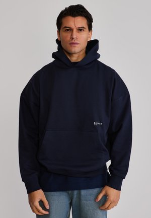 Homme portant un sweat à capuche bleu marine avec poche avant et jeans bleu clair, debout devant un fond gris uni.