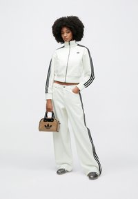 Fehér cipzáras dzseki fekete csíkokkal az ujjakon, széles szárú fehér nadrággal párosítva. A modell egy kis barna táskát tart fekete logóval.