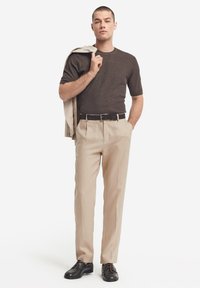 Brun ribstrikket kortærmet skjorte, beige plisserede bukser, sort bælte og sorte sko. Let jakke lagt over skulderen. Enkel, skræddersyet design.