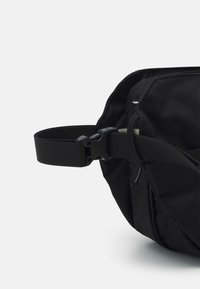 Bolsa de tela negra con un acabado texturizado. Cuenta con una correa de tejido ajustable y un bolsillo lateral con cremallera y un cierre elegante.