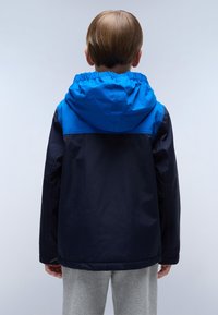 Kleurblokjack met blauwe capuchon en marineblauw lichaam. Voorzien van elastische manchetten en een verstelbare capuchon, gemaakt van een soepele, lichte stof.
