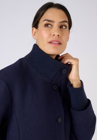 Cappotto in lana blu navy con collo alto a coste, due grandi bottoni e polsini a coste. Tessuto morbido e design sartoriale.
