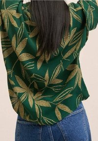 Grüne Bluse mit einem Blattmuster in Gold, mit einem entspannten Schnitt und lockeren Ärmeln, kombiniert mit blauen High-Waist-Jeans.