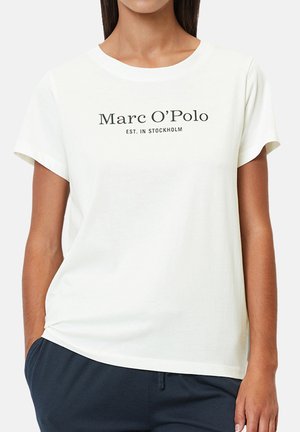 Woman wearing a white short-sleeve t-shirt with "Marc O’Polo EST. IN STOCKHOLM" printed on the front, paired with dark pants.