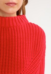 Czerwony sweter dzianinowy z wysokim prążkowanym kołnierzem, z teksturowanym wzorem i luźnym krojem na ramionach. Miękka, gruba włóczka.