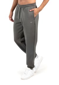 Pantalones de chándal grises con bolsillos laterales y cintura elástica. Presentan puños acanalados y un pequeño detalle de logo en la pierna. Calzan zapatillas blancas.