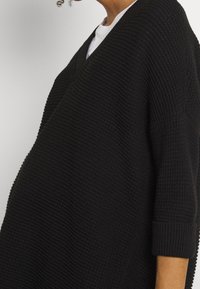 Czarny, dziergany, oversize'owy sweter z dekoltem w serek, ściągaczami na rękawach i teksturowaną powierzchnią. Rękawy o długości do połowy przedramienia, luźne i luźno opadające.