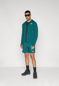 Teal athletic jacket with zip, black t-shirt, teal shorts, black sneakers, and sunglasses. Notable logo on the jacket and shorts.