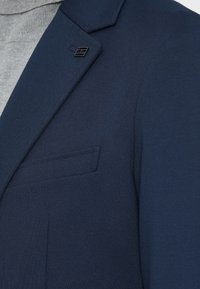 Lapel de um blazer azul-marinho com um pequeno alfinete prateado, usado sobre um suéter de gola alta cinza, mostrando a textura detalhada do tecido e as costuras.