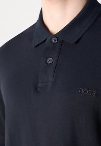 Polo bleu marine avec deux boutons fermés et logo brodé "BOSS" sur la poitrine gauche porté par une personne.