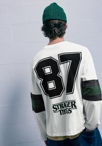 Biela košeľa s dlhým rukávom so širokými čiernymi a zelenými pruhmi, veľké "87" na chrbte a čierny nápis "STRANGER THINGS" v spodnej časti.
