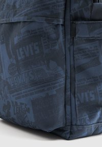 Μπλε υφασμάτινο σακίδιο πλάτης με υφή στην επιφάνεια και συνολικό μοτίβο με λογότυπα και διαφημίσεις της μάρκας Levi's σε πιο σκούρο τόνο.