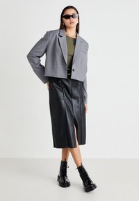 Blazer gris cintré avec un bouton unique, haut vert olive, jupe en cuir synthétique noire avec fente frontale, et bottines noires à lacets.