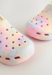 Barviti klumpi tie-dye, izdelani iz fleksibilnega materiala, z ventilacijskimi odprtinami in teksturiranimi podplati za oprijem. Prevladujeta mehki pastelni odtenki.