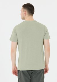 Man med kort mörkt hår och silverfärgad hoop-örhänge, iklädd en ljusgrön kortärmad skjorta och mörka byxor, vänd bort från kameran mot vit bakgrund.