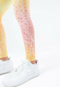 Leggings in een gradient van geel naar perzik met ronde luipaardvlekken en witte tekst. Gecombineerd met witte Nike Air Force 1 sneakers.