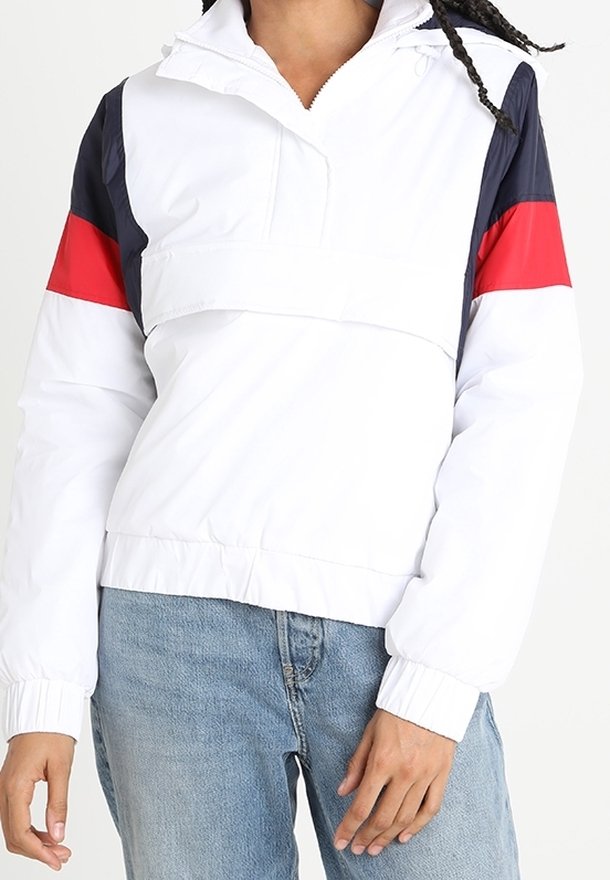 Pull blanc avec des blocs de couleurs bleu marine et rouge sur les manches, porté avec un jean bleu clair sur une personne aux cheveux tressés.