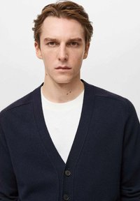 Mornarska jopica z V-izrezom, ki ima dva gumba in teksturirano blago. Nosi se čez belo srajco, prikazano z bližnjim posnetkom.