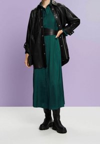 Chemise-veste oversize en cuir noir portée sur une robe midi en satin vert, cintrée à la taille avec une ceinture, accompagnée de bottines noires épaisses.