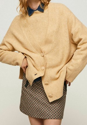 Kobieta ubrana w beżowy kardigan na guziki, na niebieskiej koszuli z kołnierzykiem oraz w kraciastą mini spódniczkę z zamykanymi kieszeniami.