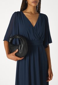 Svart läderhandväska med ett knutet handtag, i kombination med en djup marinblå klänning med V-ringning och flödande ärmar.