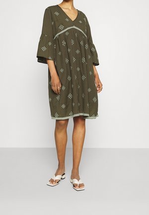 Robe vert olive avec décolleté en V, manches cloche et motifs en diamants brodés. Dentelle au niveau de la taille et de l'ourlet. Associée à des sandales blanches.