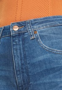 Tmavě modré džíny z denimu mají klasický pětikusový design, měděné nýty a brandovaný knoflík v pase. Světlejší vyblednutí dodává texturu.