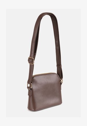 Sac bandoulière en cuir marron avec une bandoulière réglable et une fermeture éclair dorée, présenté sur un fond blanc uni.
