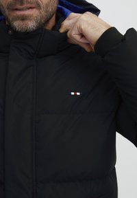 Sort pufferjakke med høj krave, der har en blå indvendig foring og et lille flerfarvet logo. Tekstureret stof med en glat finish.
