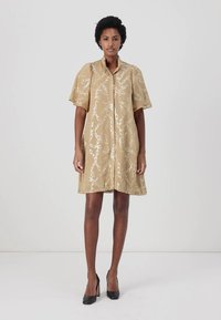 Abito beige a maniche corte con motivo a catena, chiusura con bottoni e vestibilità comoda, abbinato a tacchi alti neri. Tessuto dalla consistenza liscia.